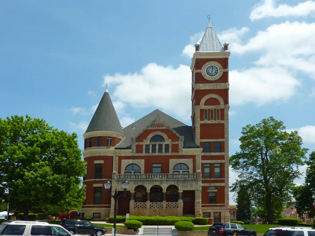Historic red brick courthouse with clock tower, arched windows, and surrounding greenery under blue sky.