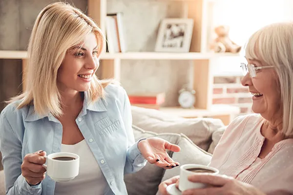 Two women, one younger and one older, smiling and conversing while holding coffee cups in a cozy living room.