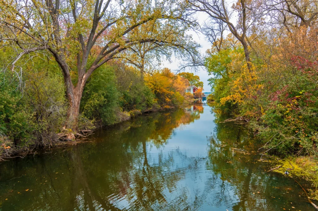 Calm river flanked by autumn trees with yellow, orange, and green foliage, and a house with a bridge in the distance.