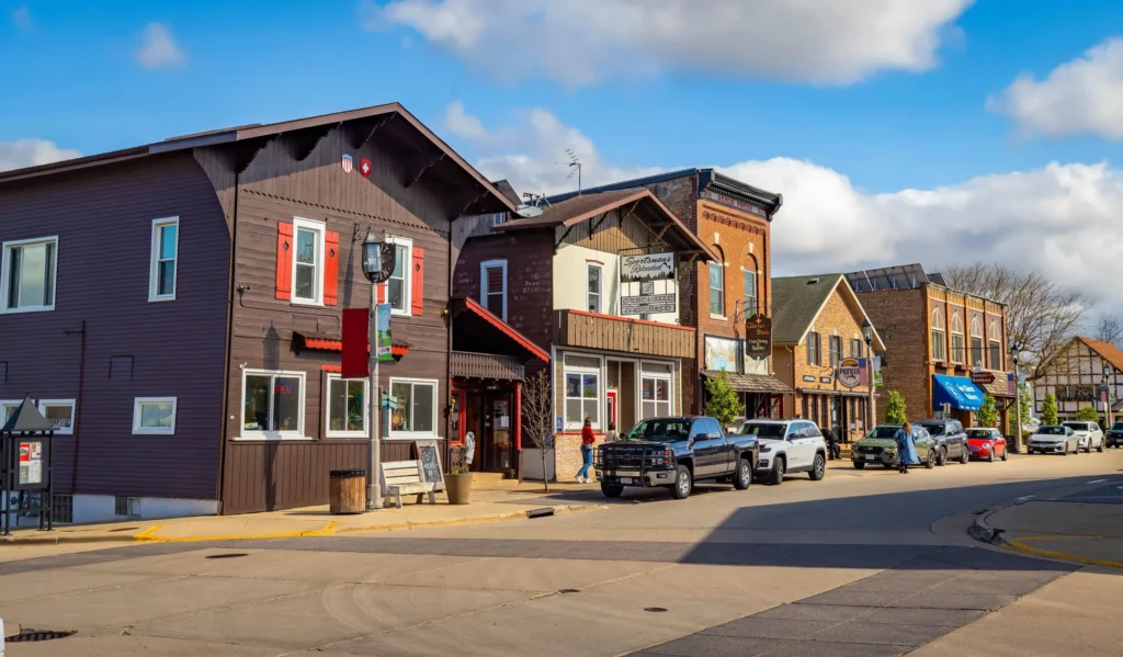 Street view of a small town with wooden and brick buildings, parked cars, and pedestrians.