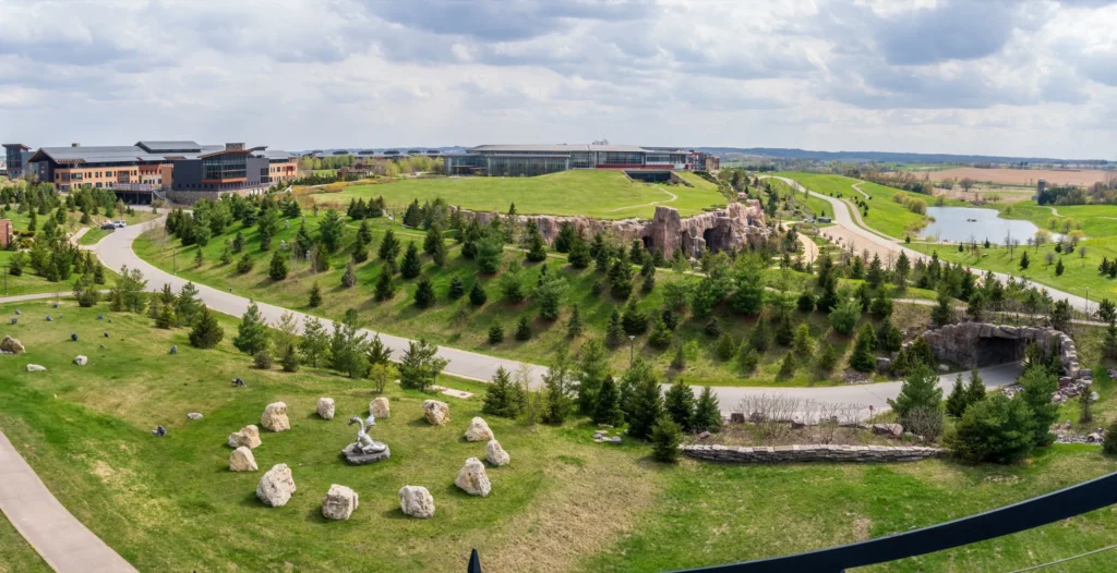 Aerial view of a green campus with modern buildings, winding roads, trees, rocks, and a pond under a cloudy sky.