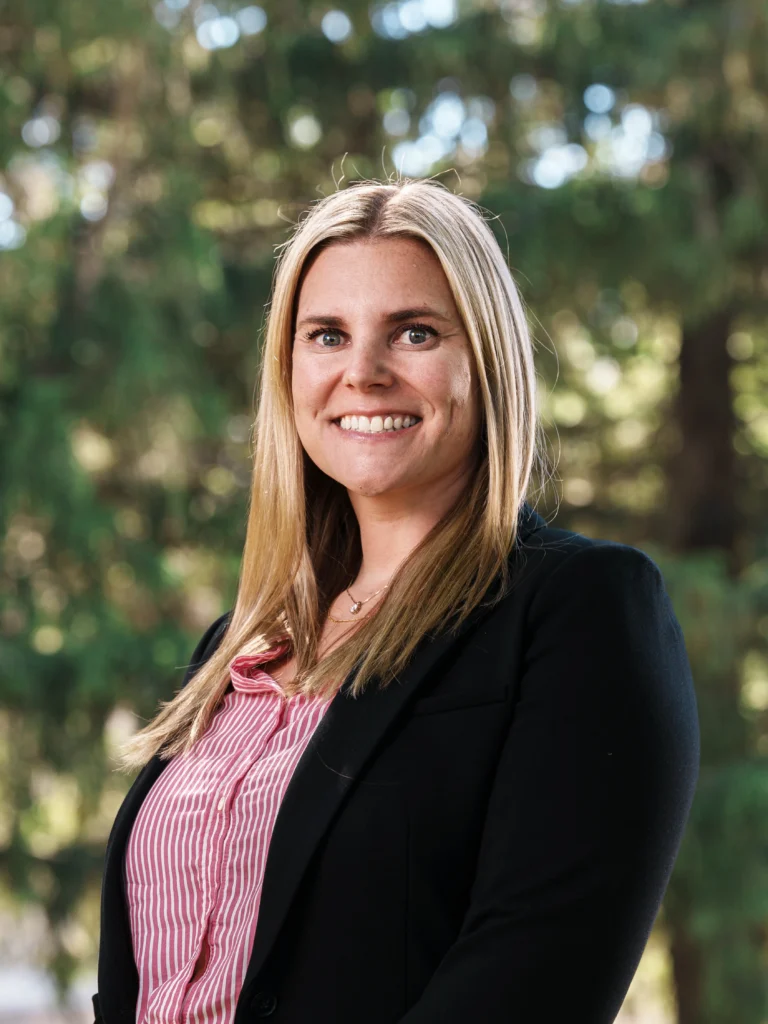 Professional portrait of a smiling blonde woman in a black blazer, outdoors with blurred greenery background.