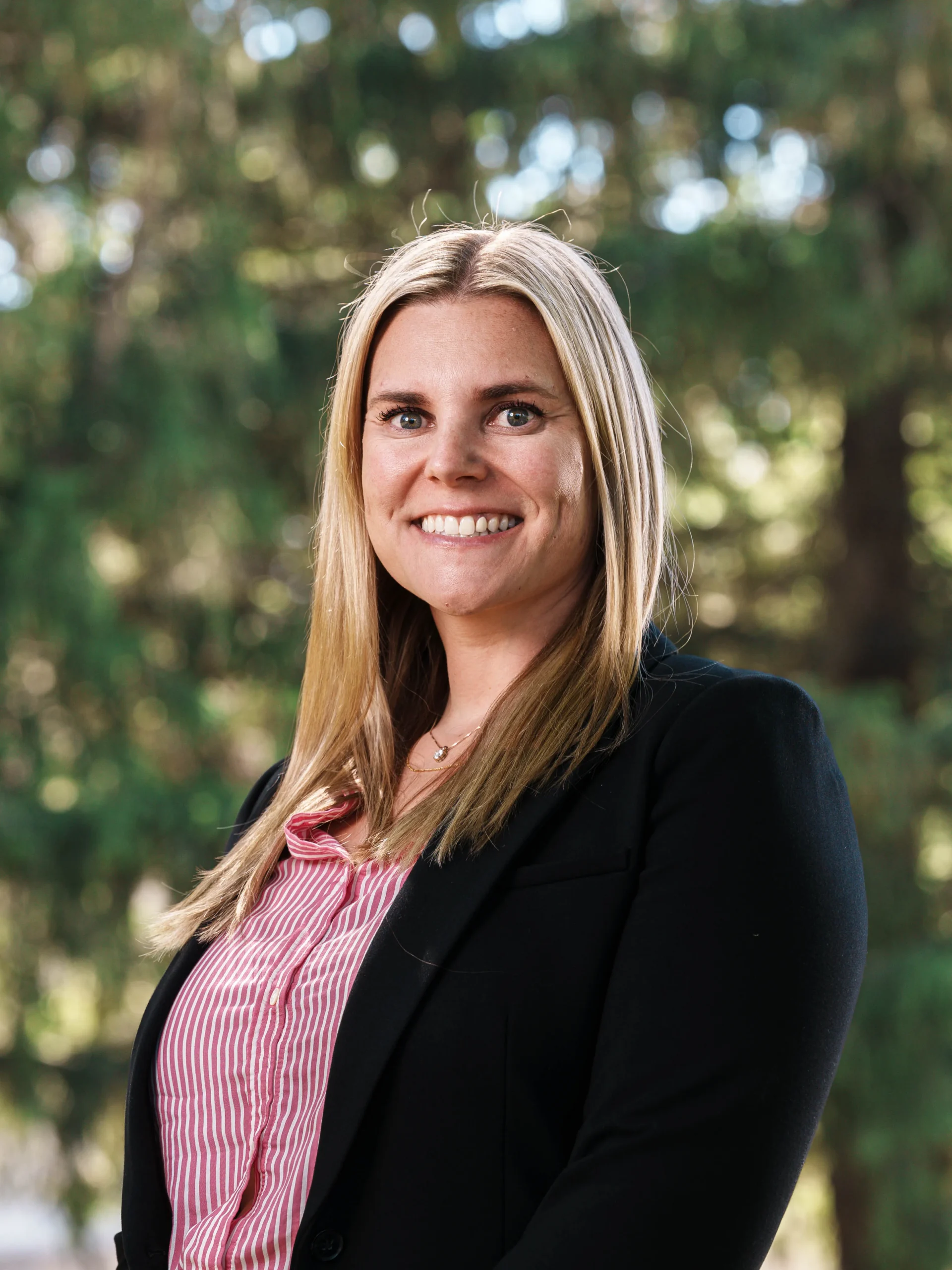 Professional portrait of a smiling blonde woman in a black blazer, outdoors with blurred greenery background.