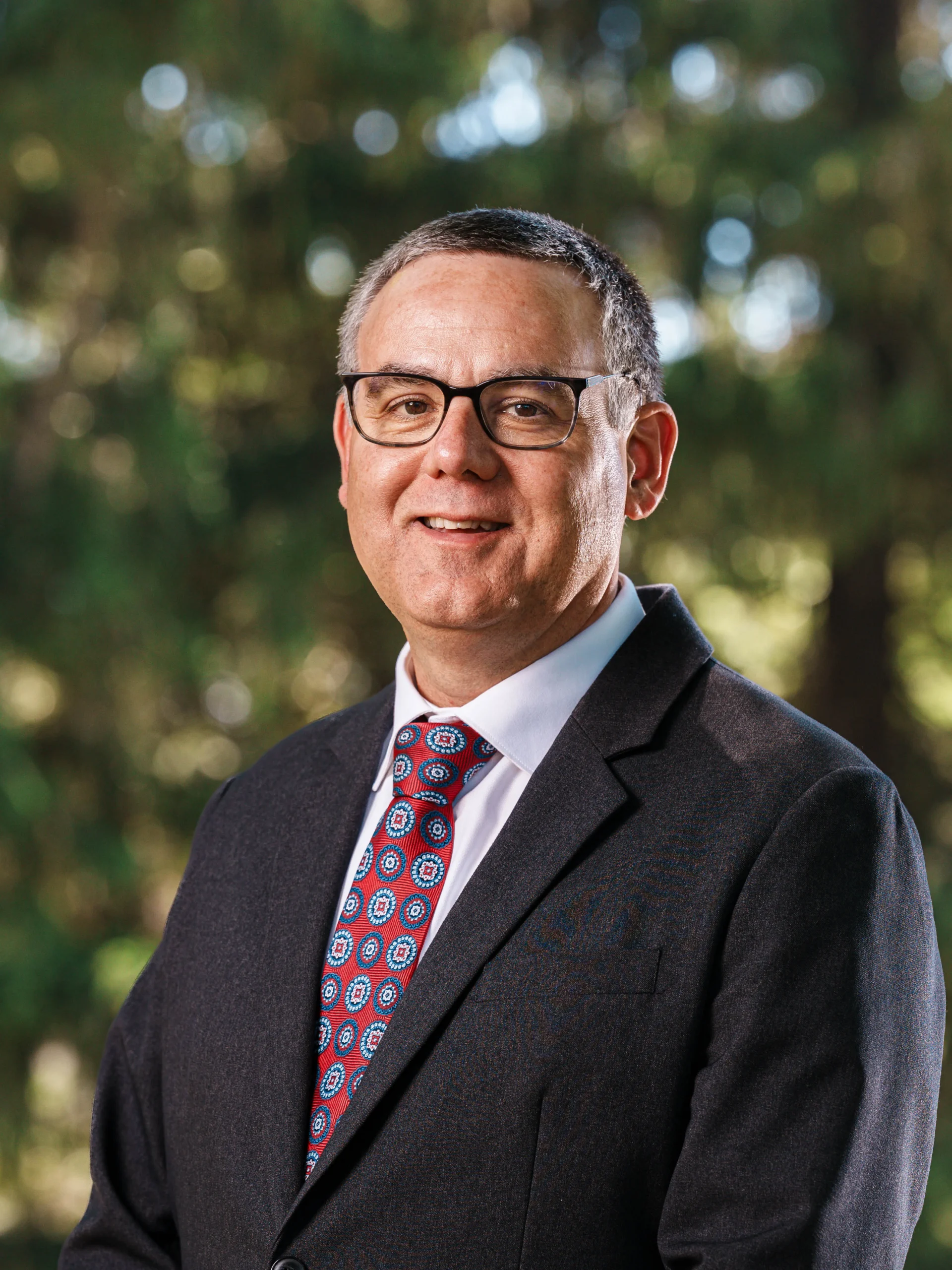 Middle-aged man with short gray hair and glasses, wearing a dark suit and patterned red tie, smiling outdoors.