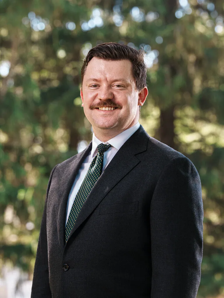 Professional portrait of a smiling man with a mustache wearing a dark suit and green tie outdoors.