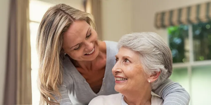 Two women, one elderly with gray hair and one younger with blonde hair, smiling warmly indoors.