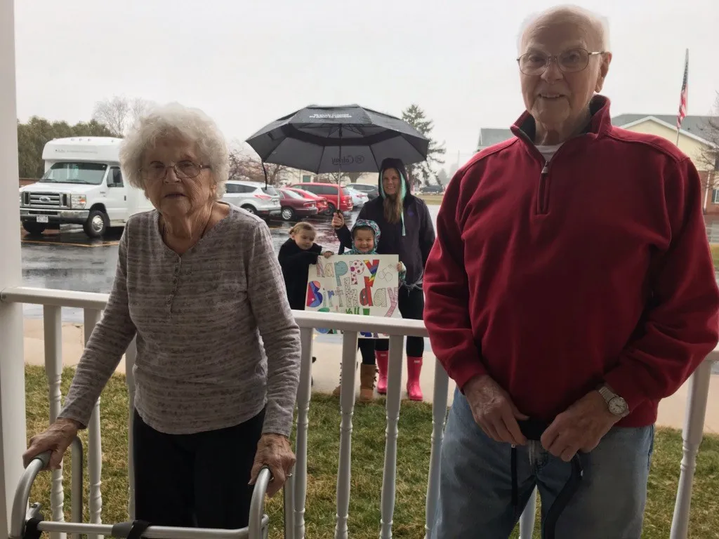 Elderly couple standing on porch with walker and umbrella, children holding colorful birthday sign in background.