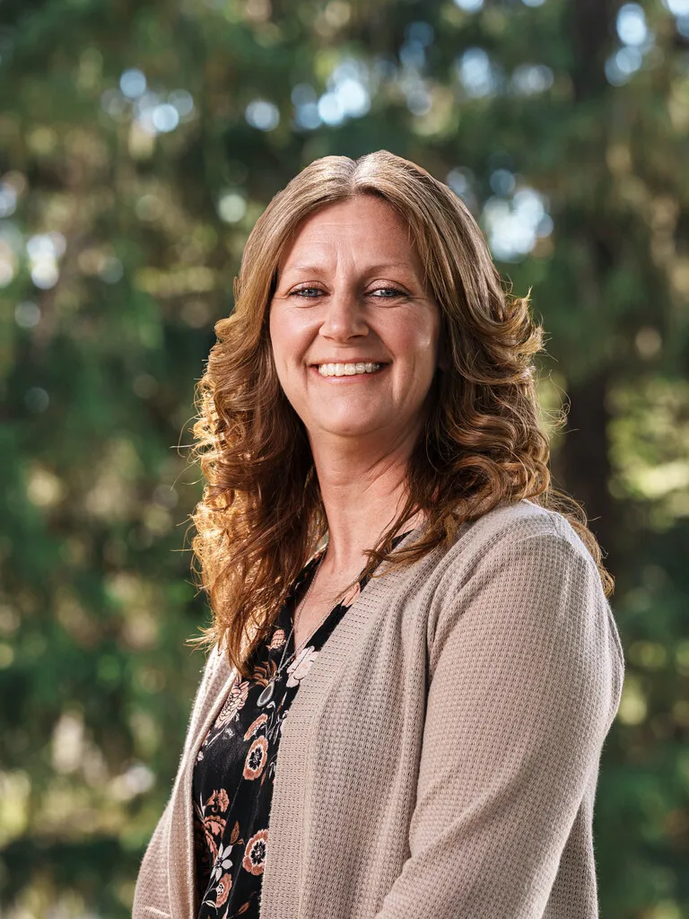 Portrait of a smiling woman with wavy light brown hair, wearing a beige cardigan and floral blouse, outdoors.