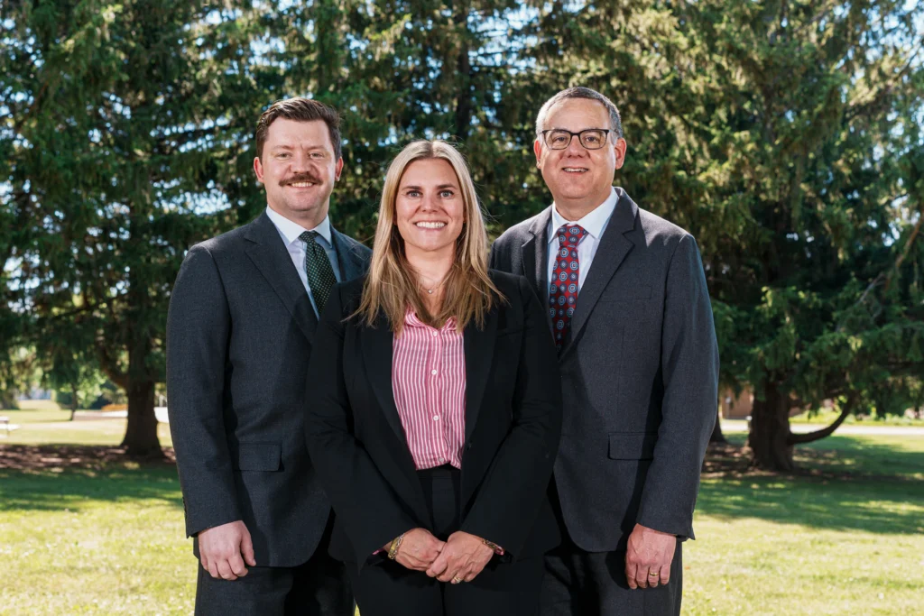Three professionally dressed individuals smiling outdoors with green trees in the background.