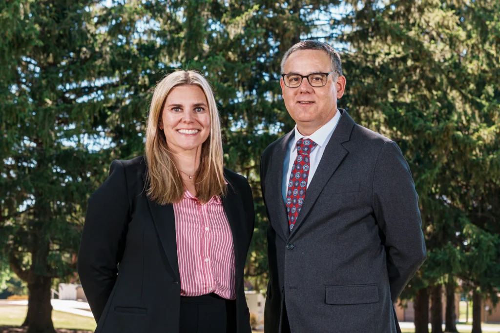 Two professionally dressed individuals, a woman and a man, standing outdoors with trees in the background.