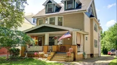 Two-story beige house with American flag on front porch and surrounding greenery.