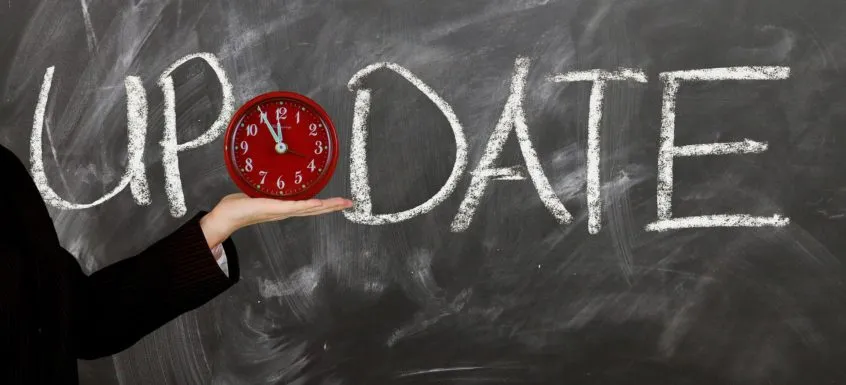 Person's hand holding a red clock showing 12:00 in front of a blackboard with "UPDATE" written in white chalk.