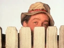 Man wearing a beige hat peeking over a white wooden fence against a plain background.